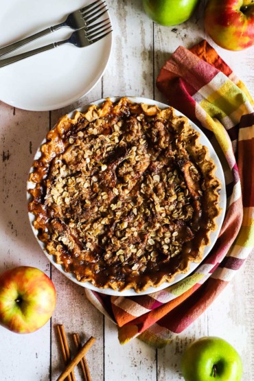 An overhead view of an unsliced caramel apple pie surrounded by whole apples and cinnamon sticks. 