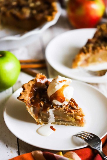 A slice of caramel apple pie on a white dessert plate topped with a scoop of vanilla ice cream and a drizzle of caramel sauce. 