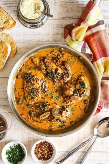 An overhead view of a large silver skillet filled with creamy Tuscan chicken with slices of toasted bread and a bowl of grated cheese nearby.