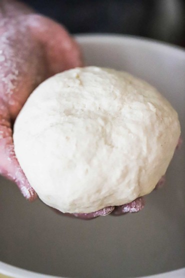 A person holding a round piece of dough over a large ceramic bowl.