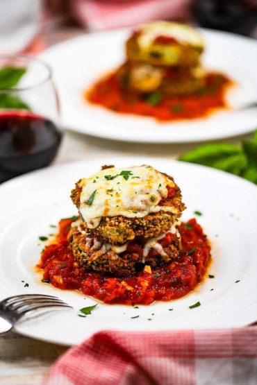 Air fryer eggplant parmesan that is two stacked breaded and fried eggplant circles topped with cheese and on a small pool of marinara sauce on a white plate. 