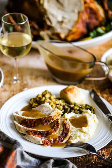 A white dinner plate filled with sliced turkey, mashed potatoes, green beans, and gravy next to a glass of white wine and a platter of cooked turkey. 