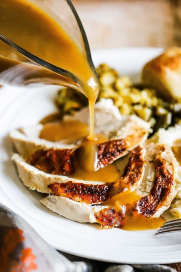  A glass gravy boat being used to pour turkey gravy over sliced fried turkey on a white dinner plate. 