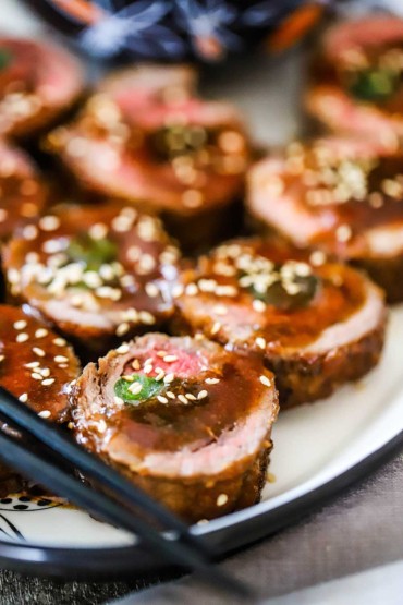 A close-up view of slices of beef negimaki on a platter topped with a brown sauce and sesame seeds.