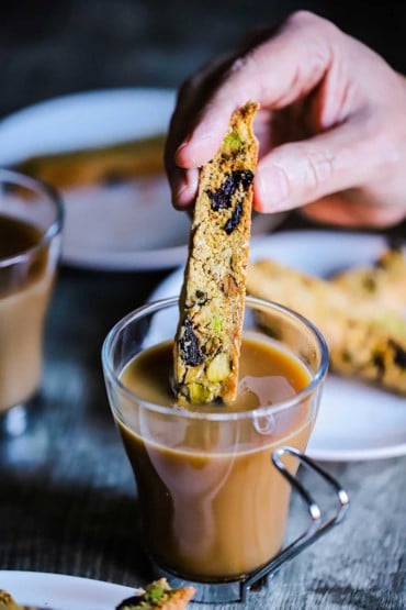 A person dunking a biscotti with pistachios and dried cherries into a glass mug filled with creamed coffee.