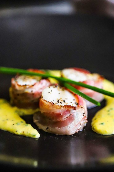 A close-up view of sea scallops with pancetta wrapped around them sitting on a black plate with an egg and chive sauce surrounding them.