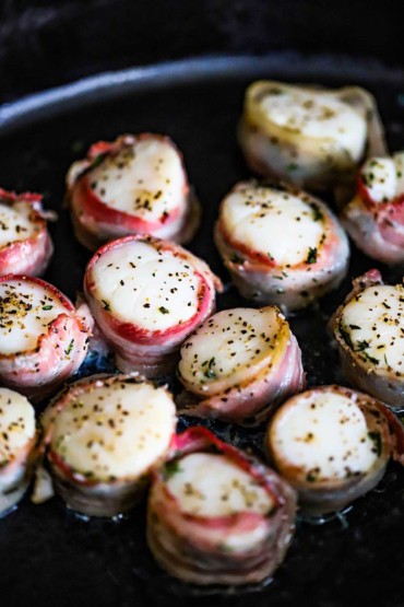 A black oval baking dish filled with sea scallops that have been baked with thin strips of pancetta wrapped around them.