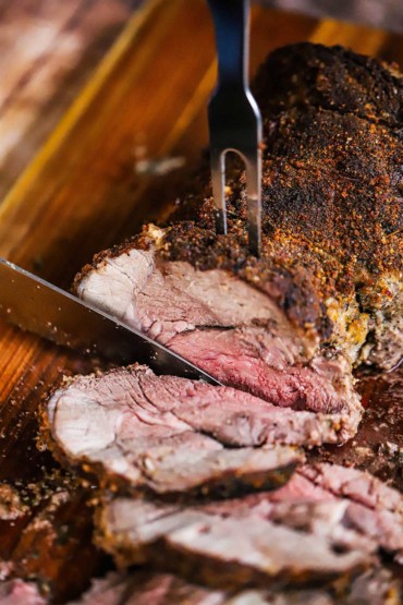 A meat carving fork stuck into a boneless leg of lamb with a carving knife separating slices from it on a cutting board.