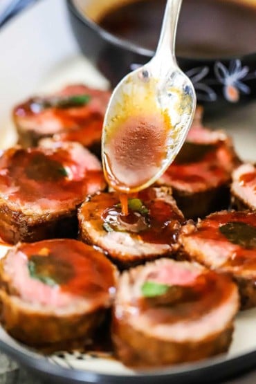 A spoon being used to drizzle a brown Asian sauce over the tops of sliced beef negimaki rolls on a platter.