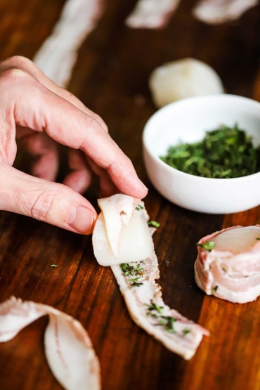 A person wrapping a thin slice of pancetta around a sea scallop with a small bowl of freshly chopped thyme nearby.