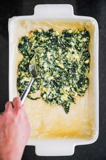 A person using a large spoon to add a spinach and feta cheese mixture into a baking dish that has been lined with buttered sheets of phyllo.