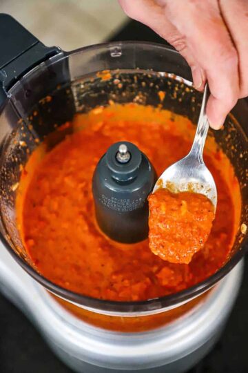 A person holding up a spoonful of homemade romesco sauce from a food processor.