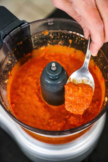 A person holding up a spoonful of homemade romesco sauce from a food processor.