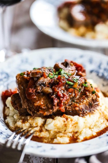 A dinner bowl filled with a pile of mashed potatoes that is topped with two sliced of Italian pot roast with a chunky tomato sauce poured over the top.