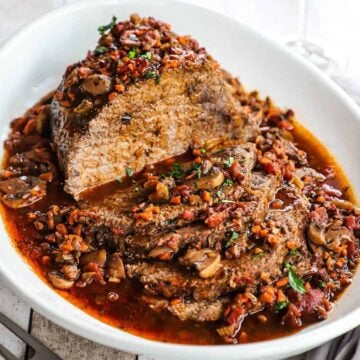 An Italian pot roast that has been sliced and is sitting in a large oval platter with a tomato and mushroom sauce all over it.