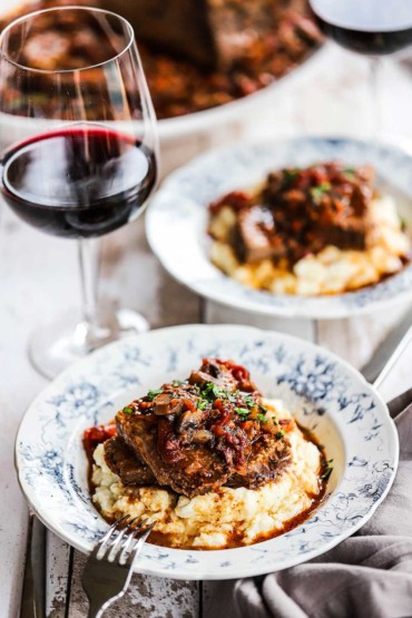 Two antique dinner bowls filled with mashed potatoes and topped with slices of Italian pot roast all sitting next to a glass of red wine.