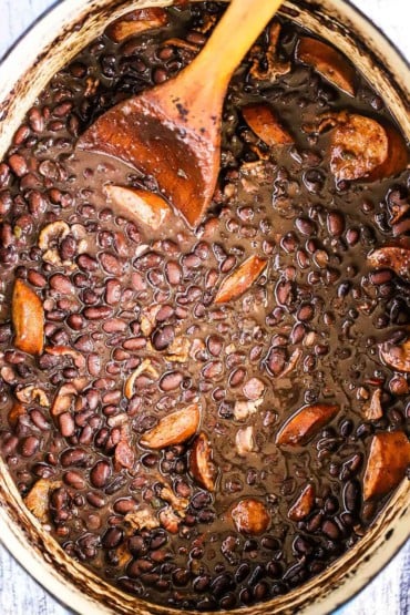 An overhead view a large oval Dutch oven that is filled with black bean and andouille sausage with a wooden spoon stuck into it.