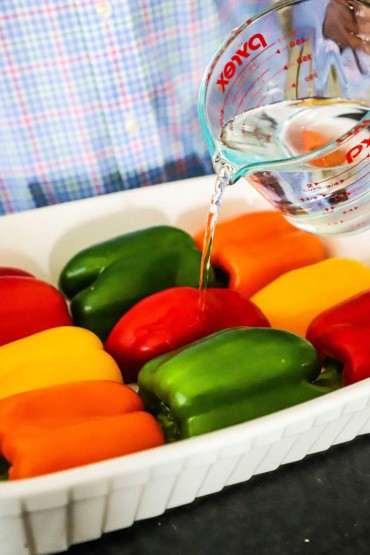 A person pouring water from a measuring cup into a white baking dish filled with halved bell peppers sitting cut-side down.