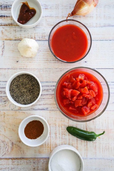 A collection of bowls holding a chipotle pepper, tomato sauce, diced, tomatoes, salt, pepper, chili powder, and onion and pepper nearby.