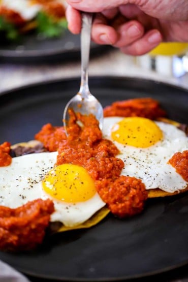 A person using a small spoon to add a layer of ranchero sauce over two fried eggs that are sitting on two lightly fried corn tortillas on a black plate.