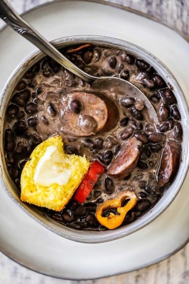 An overhead view of a soup bowl filled with black bean soup with andouille and topped with a piece of buttered cornbread and slices of peppers.
