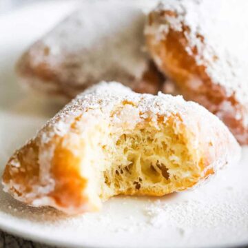 A close-up view of a half-eaten beignet covered in powdered sugar sitting on a white dessert plate.