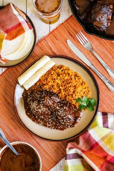An overhead view of a plate filled with chicken mole and Mexican rice next to rolled corn tortillas.