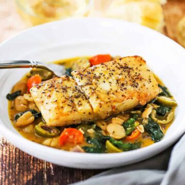 A close-up view of a seared fillet of halibut sitting on top of sautéed spinach, tomatoes, olive, and beans all in a savory sauce.