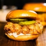 A close-up side view of a fried chicken sandwich with pickles and remoulade between toasted brioche buns.