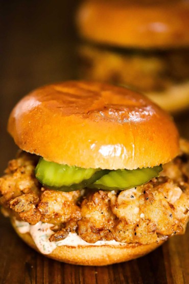 A crispy fried chicken sandwich sitting on a cutting board loaded with pickles and a remoulade sauce.
