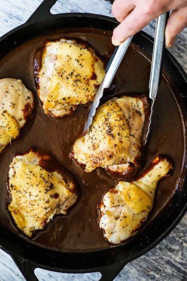 A person using a pair of metal tongs to nestle seared chicken pieces into a skillet filled with mole poblano sauce.