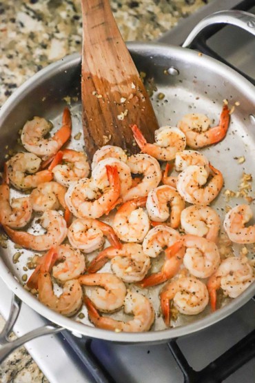 An overhead view of pink seared shrimp in a large stainless steel skillet with a wooden spatula off the side.