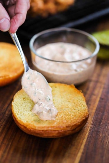 A person using a spoon to smear a Cajun remoulade over the bottom half of a toasted brioche bun.