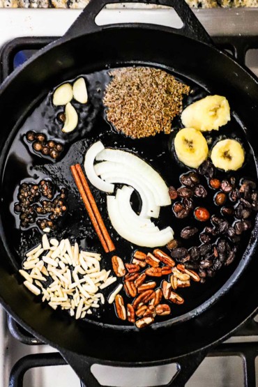 An overhead view of a cast-iron skillet that is filled with small portions of nuts, fruits, seasonings, onion, and a cinnamon stick all sizzling in a small layer of oil.