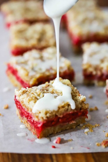 A vanilla glaze is being drizzled from a spoon onto a square raspberry linzer bar sitting next to other linzer bars on parchment paper.