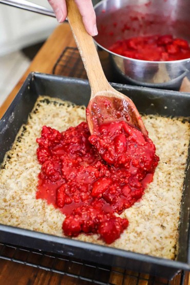 A person transferring a raspberry filling over the top of a partially baked shortbread crust in a metal square baking pan.