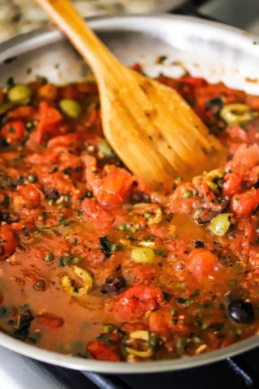 A close-up view of a large stainless steal skillet filled with a puttanesca sauce with a wooden spatula inserted.