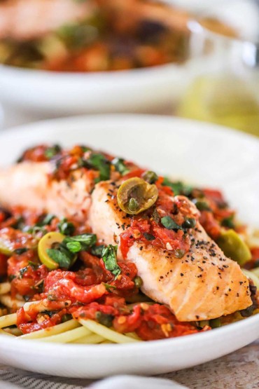 A close-up view of a shallow white pasta bowl filled with salmon puttanesca sitting on a bed of pasta.