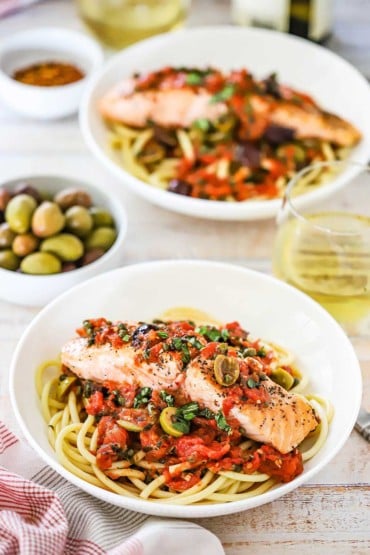 An overhead view of two shallow pasta bowls each filled with a bed of bucatini pasta topped with salmon and a puttanesca sauce.