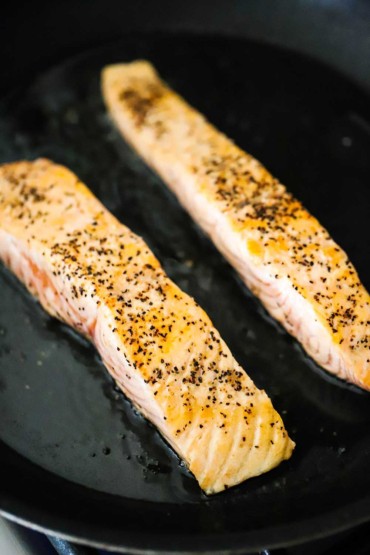Two thin strips of salmon fillets being seared in olive oil in a black non-stick skillet.