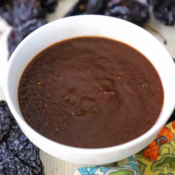 A white bowl filled a brown ancho chili sauce that is sitting next to dried ancho chilils and a festive colorful napkin.