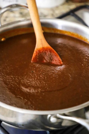 A wooden spoon inserted into the middle of a stainless steel skillet filled with a smooth brown enchilada sauce.