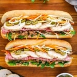 An overhead view of two side-by-side banh mi sandwiches that are stuffed with cilantro, cold cuts, sliced pork roll, carrots, daikon, and pickled jalapeños.
