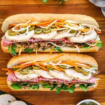 An overhead view of two side-by-side banh mi sandwiches that are stuffed with cilantro, cold cuts, sliced pork roll, carrots, daikon, and pickled jalapeños.
