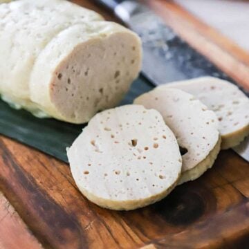 Three slices of cha lua sitting on a cutting board lined with a banana leaf and a loaf of the cha lua nearby.