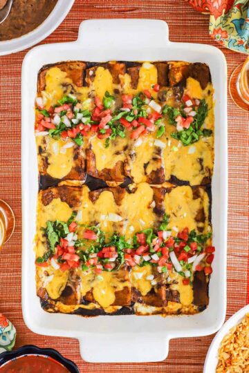 An overhead view of a white 9 by 13 baking dish filled with cheese enchiladas in ancho sauce topped with shredded lettuce and pico de Gallo.