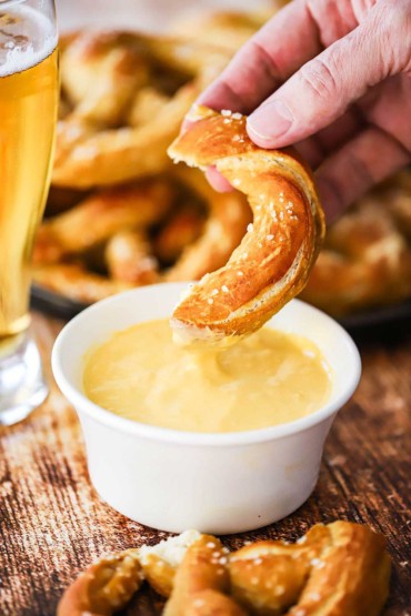 A person raising a portion of a pretzel that has been dipped into a bowl of cheese sauce.