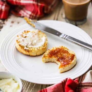 Two homemade English muffin halves covered with butter and peach jam with one that has a bite taken out of it.