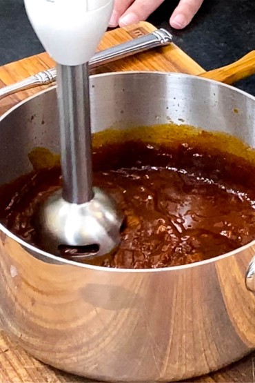 A person using an immersion blender to purée a curry tomato sauce in a silver saucepan.