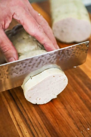 A person using a Japanese-style knife to cut a slice of cooked cha lua on a cutting board.